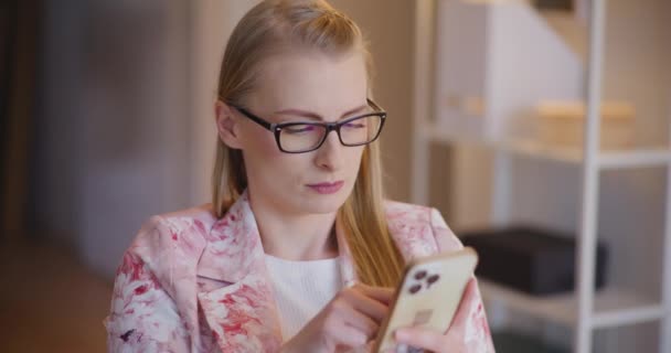 Молода блондинка-підприємець прокручує мобільний телефон, сидячи в офісі - Кадри, відео