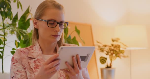 Jovem empresária loira confiante usando blazer floral usando tablet digital enquanto navega na internet no escritório - Filmagem, Vídeo
