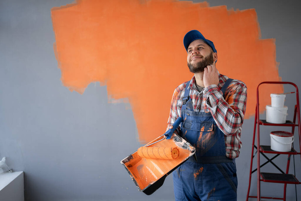 Malermann mit Farbroller und Tablett in der Hand steht zweifelnd und mit verwirrtem Gesichtsausdruck neben Malwand. Kopierraum - Foto, Bild