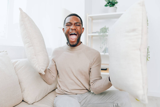 Relaxed African American man sitting on a comfortable sofa in a modern living room He is smiling and holding a pillow, enjoying his weekend at home The background is stylish and the atmosphere is - Photo, Image