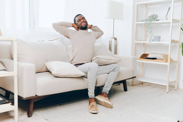 Relaxado homem afro-americano sentado em um sofá confortável em casa, sorrindo e desfrutando de seu fim de semana Ele segura um travesseiro enquanto pensa, com uma sala de estar moderna como pano de fundo Este jovem e - Foto, Imagem