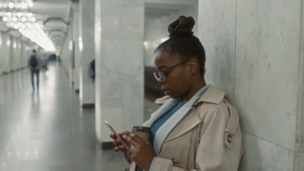 30 'lu yaşlarda gözlüklü, metro istasyonunda trenini beklerken telefondaki mesajları kontrol eden ve elinde tek kullanımlık kahve fincanı tutan siyahi bir kadının görüntülerini sandığa koy. - Video, Çekim