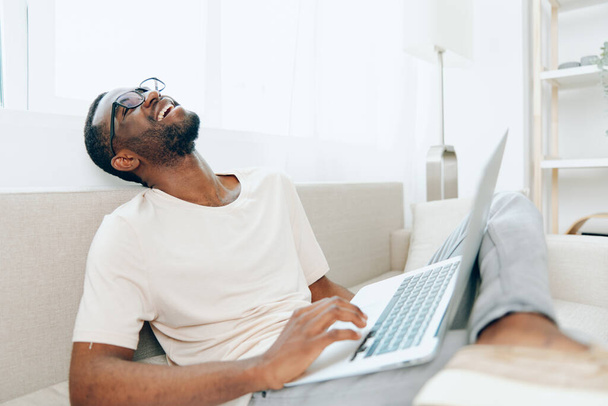 Joven afroamericano freelancer trabajando en un ordenador portátil y escribiendo felizmente mientras está sentado en un sofá moderno en una acogedora sala de estar Con un ambiente informal y relajado, abraza los beneficios de la educación - Foto, Imagen