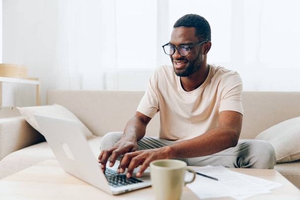Hymyilevä afroamerikkalainen mies työskentelee kannettavalla tietokoneella sohvalla modernissa kodissa Kuvassa on nuori tuhatvuotinen freelancer, yllään rento vaatteita, mukavasti istuu mustalla sohvalla olohuoneessaan. - Valokuva, kuva