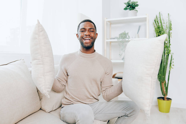 Relajado hombre afroamericano sentado en un cómodo sofá en su moderna sala de estar Él está sonriendo y sosteniendo una almohada mientras piensa y disfruta de un fin de semana tranquilo en casa El fondo muestra - Foto, Imagen