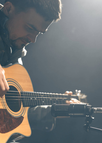 Ein männlicher Gitarrist spielt eine akustische Gitarre mit Mikrofon auf dunklem Hintergrund in einem Tonstudio oder Konzertsaal. - Foto, Bild