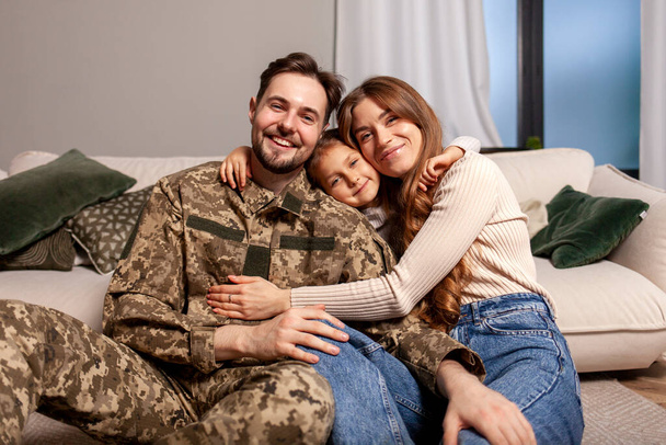Ukraiński żołnierz armii w mundurze kamuflażu wrócił do domu do rodziny, kadeci wojskowi siedzi na kanapie z żoną i córką i uśmiecha się, dziecko przytula weteranów ojciec - Zdjęcie, obraz