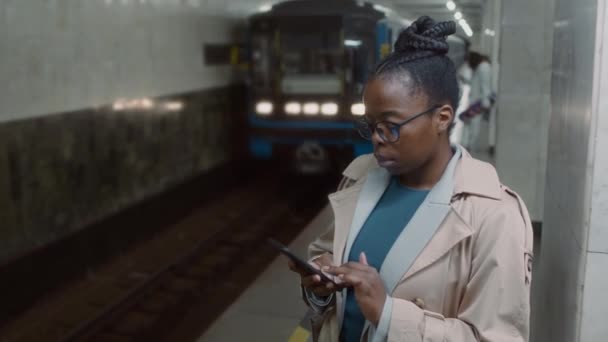 Imágenes secundarias de la señora afroamericana escribiendo mensajes en el teléfono en la plataforma del metro y su tren eléctrico azul finalmente llegando a la estación - Imágenes, Vídeo