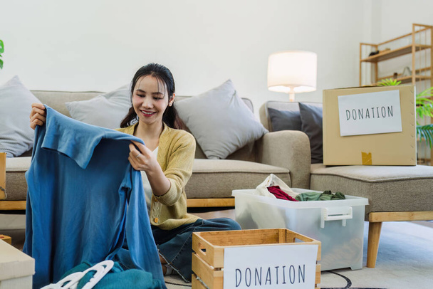 Asyalı genç bir kadın evde giysilerini topluyor, ikinci el kıyafetlerle bağış kutusuna bir şeyler koyuyor.. - Fotoğraf, Görsel