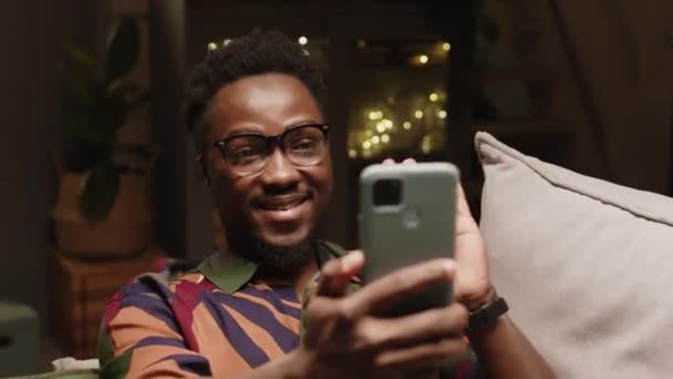 Περιμένετε ένα νεαρό θετικό μαύρο άνδρα που φοράει patterned hipster πουκάμισο και γυαλιά χαιρετισμό και μιλάμε ενώ έχουν βιντεοκλήση στο smartphone στο σπίτι το βράδυ - Πλάνα, βίντεο