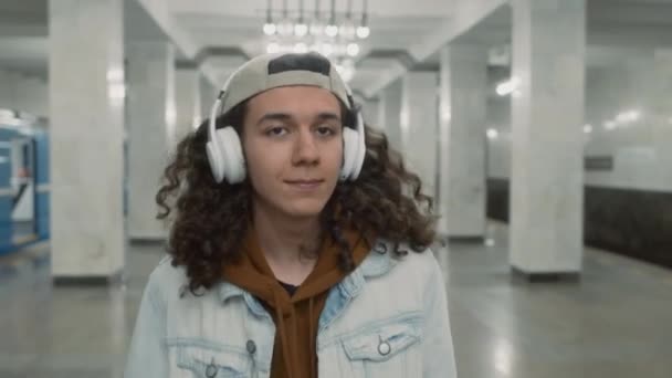 Het opsporen van borst-tot-borst beelden van positieve blanke knappe man met krullend haar lopen alleen langs verlichte metro lobby en luisteren naar muziek in koptelefoon - Video