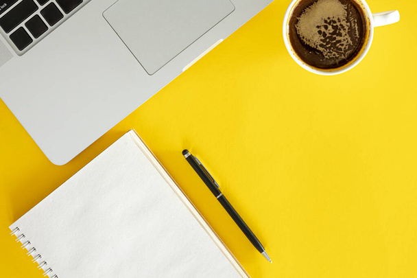 Kopje koffie, notitieblok en laptop op gele achtergrond, plat gelegd. Minimale kantoorachtergrond, kopieerruimte. - Foto, afbeelding