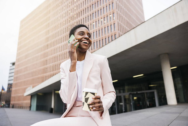 Joven hermosa mujer de negocios sonriente sosteniendo taza reutilizable y hablando en un teléfono verde durante una pausa de café en la calle en la ciudad. - Foto, Imagen