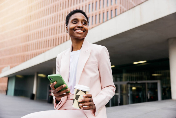 Mladá krásná usměvavá podnikatelka drží opakovaně použitelný hrnek a pomocí zeleného telefonu během přestávky na kávu v ulici ve městě. - Fotografie, Obrázek
