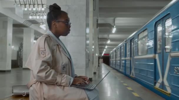 Imágenes laterales de una trabajadora independiente negra sentada en un banco subterráneo y trabajando en un portátil mientras espera otro tren - Imágenes, Vídeo