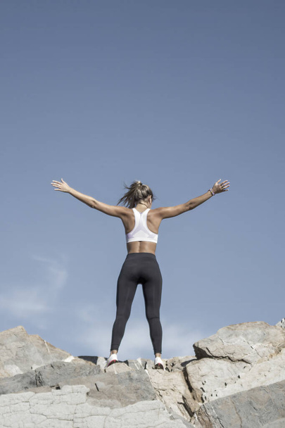 blonde vrouw in sportkleding verhogen van haar armen gelukkig op de top van een aantal rotsen - Foto, afbeelding