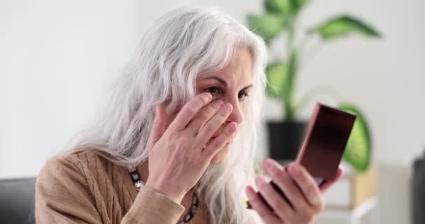 Oudere vrouw met grijs haar kijken naar rimpels en zakken onder de ogen in spiegel thuis 4k film slow motion. Leeftijdsgebonden veranderingen in gezichtshuidconcept - Video