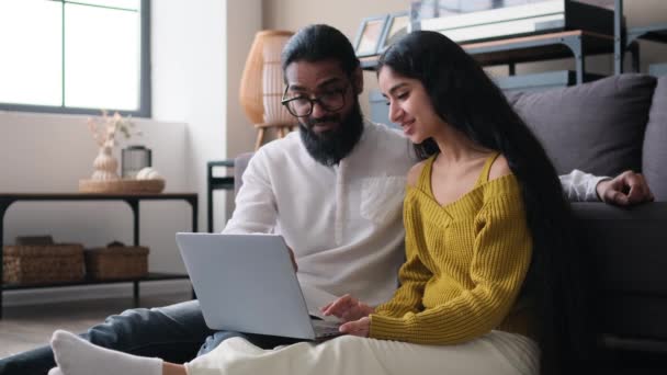 Lachende Indiase vrouw en echtgenoot gebruiken laptop om samen online te ontspannen, terwijl ze thuis op de vloer op de woonkamer zitten. Huishoudelijke vrijetijdsbesteding. - Video