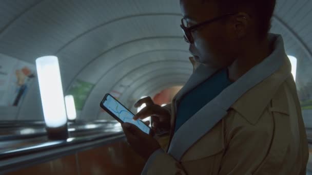 Imágenes laterales del cofre de la dama afroamericana en gabardina mirando el mapa de tránsito del metro en la pantalla del teléfono mientras desciende por las escaleras mecánicas a la estación de metro - Imágenes, Vídeo