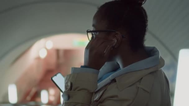 Imágenes laterales del cofre de la señora negra en gafas que se ponen auriculares y usan el teléfono móvil mientras descienden escaleras mecánicas a la estación de metro - Imágenes, Vídeo
