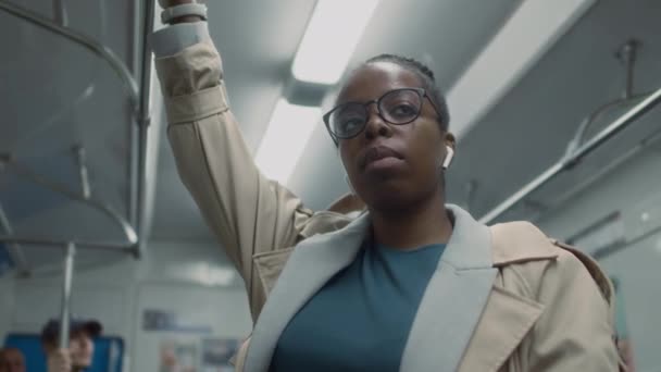 Alçak açılı göğüs görüntüsü, gözlüklü ve kulaklıklı siyahi bir kadının metro trenine binerken ayakta durduğunu gösteriyor. - Video, Çekim