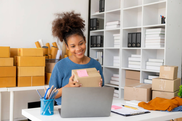 Startup, small business, African woman business owner using a smartphone or laptop to receive and verify online orders in order to prepare boxes of packed products for delivery. SME business concept - Фото, зображення