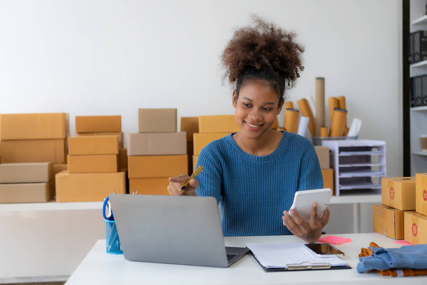 Startup, small business, African woman business owner using a smartphone or laptop to receive and verify online orders in order to prepare boxes of packed products for delivery. SME business concept - Foto, Imagem