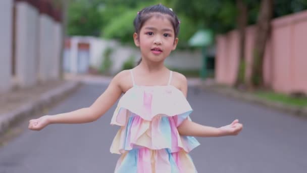 Porträt eines attraktiven kleinen asiatischen Mädchens, das lächelt und Kamera schaut und mit der Hand winkt, fröhlich lächelndes Kind, das in die Kamera schaut - Nahaufnahme, draußen. Freudiges Freizeitkonzept für die Natur. - Filmmaterial, Video