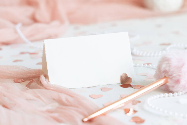Vaakasuora taitettu kortti lähellä vaaleanpunainen koristeet, kynä, sydämet ja silkkinauhat valkoisella pöydällä lähikuva. Romanttinen pöytä paikka mockup häät, Ystävänpäivä, Kevät tai Äitienpäivä paperitavarat - Valokuva, kuva