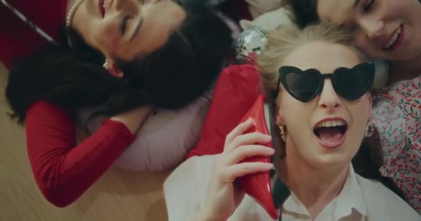 Verspielte modische junge Frau mit Sonnenbrille, Telefon in der Hand, während sie am Galentinentag bei Freundinnen liegt - Filmmaterial, Video