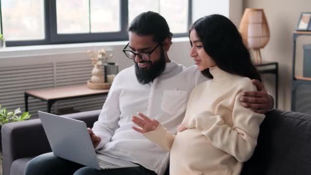Young Indian pregnant wife and husband using laptop on sofa in living room. Preparing for childbirth, doing online shopping, enjoying pregnancy time and baby expecting . - Footage, Video