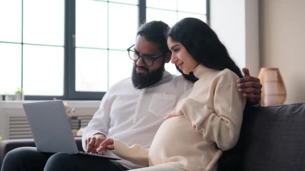 Ystävällinen intialainen perhe, vaimo ja aviomies, odottaa vauva käyttää kannettavaa tietokonetta verkkokauppaan, valmistautumiseen synnytykseen, rentoutumiseen kotona sohvalla.. - Materiaali, video