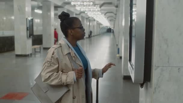 Boş yeraltı koridorunda dijital ekran kullanarak metro transit haritasını kontrol eden siyah kadın gezginin yan görüntüleri. - Video, Çekim