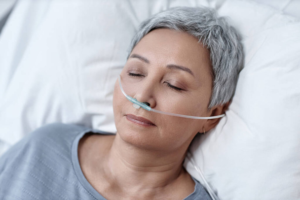 Высокий угол обзора бессознательной зрелой женщины, искусственно дышащей через трубку, лежащую на кровати в палате - Фото, изображение