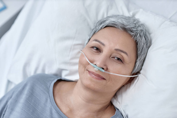 Высокий угол обзора зрелой женщины, смотрящей на камеру, лежащую на кровати с кислородной трубкой - Фото, изображение