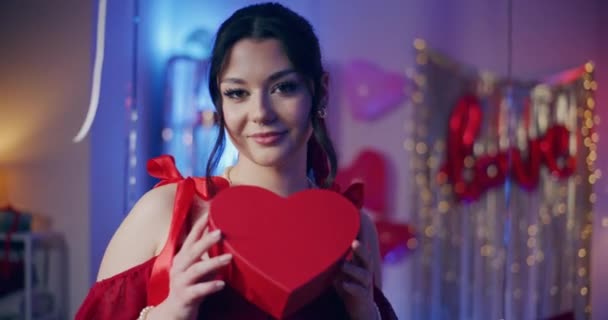 Sonriente joven atractiva sosteniendo caja de regalo en forma de corazón en casa durante la celebración del día de Galentines - Metraje, vídeo