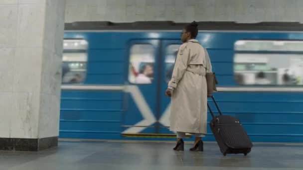 Imágenes traseras de un viajero femenino de moda con una maleta negra parada sola en la plataforma vacía del metro y mirando cómo salir del tren de tránsito del metro esperando a otro durante el viaje - Imágenes, Vídeo