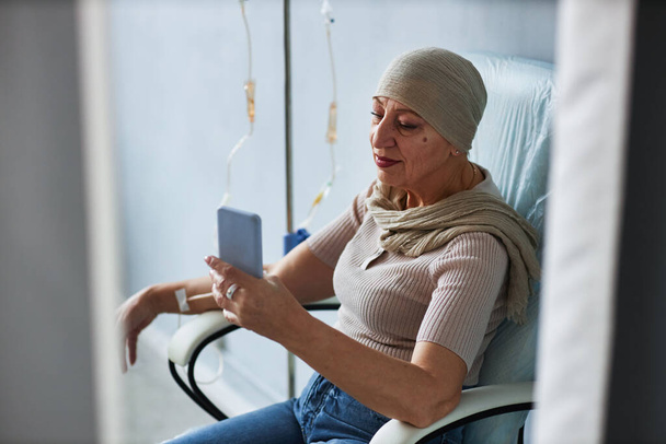 Портрет старшої жінки, що сидить у зручному кріслі з IV крапельницею та використовує смартфон - Фото, зображення