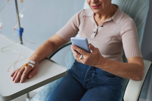 Minimal closeup of mature woman using smartphone during IV drip treatment, copy space - Photo, Image