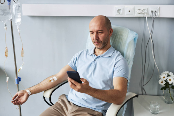 Portrait grand angle d'un homme adulte utilisant un smartphone pendant un traitement IV goutte à goutte en clinique - Photo, image
