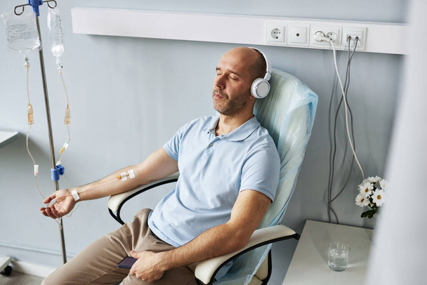 Portrait en grand angle d'un homme adulte portant un casque et se relaxant pendant un traitement IV goutte à goutte à la clinique - Photo, image