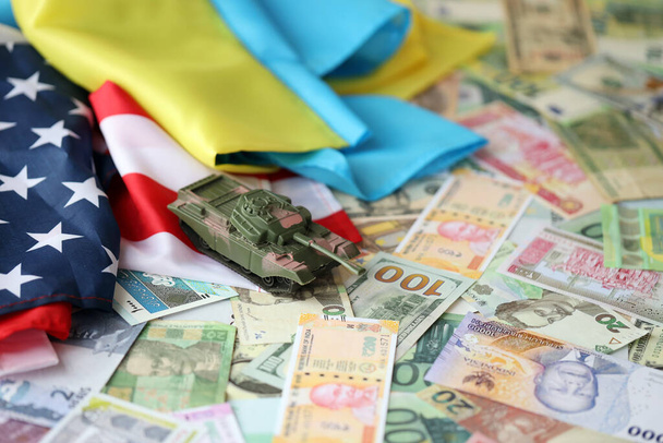 Tanque de juguete en bandera estadounidense y bandera ucraniana en muchos billetes de diferentes monedas. Antecedentes de financiación de la guerra y apoyo militar entre Estados Unidos y Ucrania - Foto, Imagen