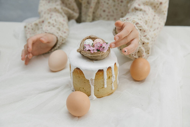 Osterkuchen mit weißer Glasur und einem dekorativen Vogelnest mit Eiern und Blumen in Kinderhänden. Kind hält Osterkuchen in der Hand. - Foto, Bild