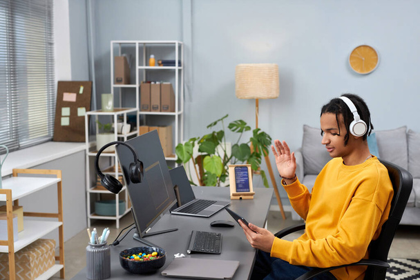 Retrato de vista lateral del joven negro como desarrollador de software usando chat de video en el lugar de trabajo y saludando - Foto, imagen