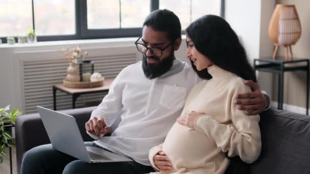 Indian husband and pregnant wife use laptop for online shopping while sitting on sofa in living room. Preparation for a child, anticipation of childbirth. - Footage, Video