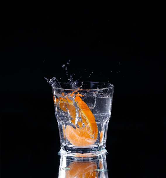 Slice of lemon splashing into a glass of water with a spray of water droplets in motion suspended in the air above the glass on a dark background. - Foto, immagini