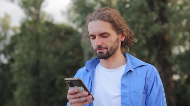 Portrait d'un homme caucasien barbu portant une chemise bleu clair tenant un smartphone moderne noir tout en se tenant debout à l'extérieur au milieu d'arbres verts. Homme utilisant gadget numérique pour naviguer dans les médias sociaux sur la rue. - Séquence, vidéo