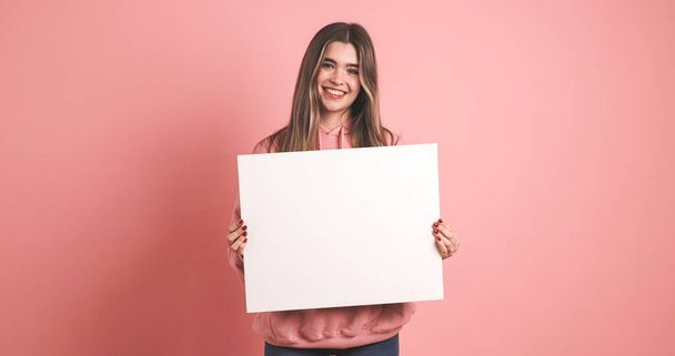 Šťastná mladá modelka dívá na kameru, zatímco drží prázdný bílý štítek pro reklamní účely na růžovém pozadí - Fotografie, Obrázek
