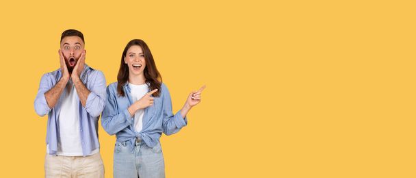 Shocked young man with open-mouthed expression and joyful woman pointing to the side at free space, both standing against vibrant yellow background, panorama - Photo, Image
