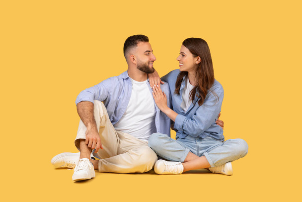 Happy man and woman sit cross-legged, facing each other in casual attire, sharing tender, joyous moment with warm embrace and loving gazes - Photo, Image